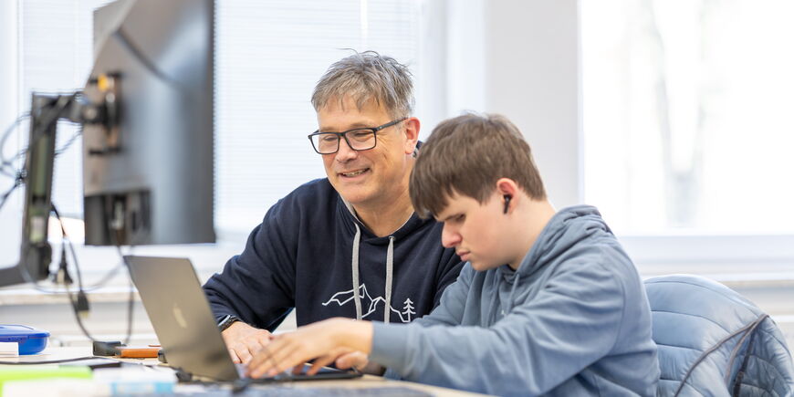 Eine Lehrkraft und ein Auszubildener sitzen an einem Laptop, der Azubi arbeitet mit einen Screenreader .