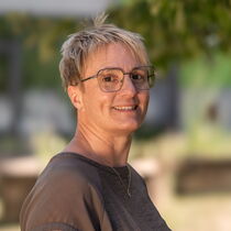 Portraitfoto Christiane Rüger - Sie trägt ein braunes Oberteil und eine Brille und hat kurzes blondes Haar.