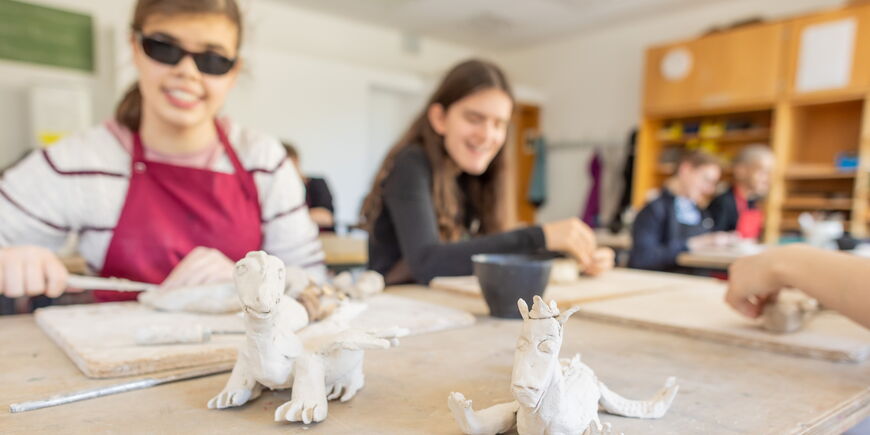 Im Vordergrund stehen zwei getöpferte Drachen, im Hintergrund arbeiten Schüler*innen an Ton.