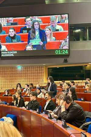 Die Autorin ist zu sehen wie sie im Plenarsaal des Europarats steht, und eine Rede hält. Im Saal hängt ein großer Bildschirm, der die Rednerin vergrößert zeigt. Am unteren Rand des Bildschirms links steht „Maike Elsasser – Allemagne“ und rechts unten auf