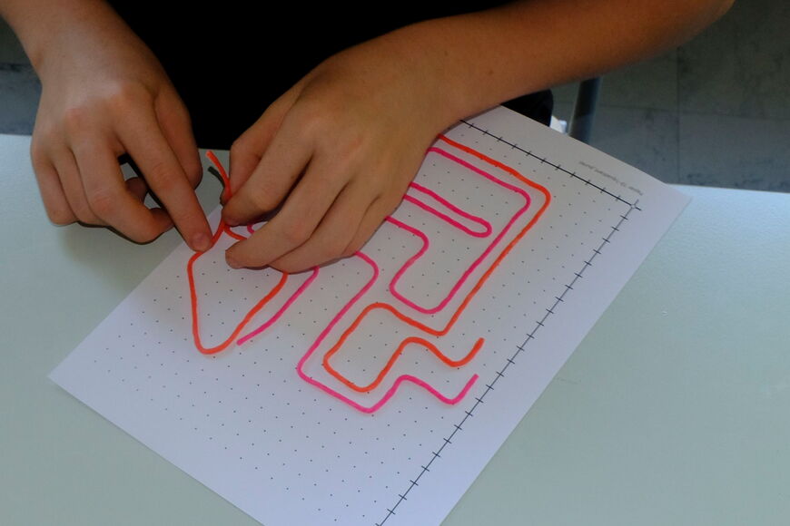 Ein Schüler sitzt an einem Tisch, darauf ein Blatt mit einem Koordinatensystem, erster Quadrant. Die Fläche des Quadranten ist mit einem Punktraster ausgefüllt. Der Schüler legt mit zwei Wikki-Sticks ein Labyrinth. Davor liegt ein weiteres Blatt, auf dem
