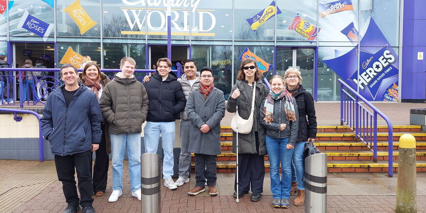 Die Gruppe Schüler*innen und Lehrer*innen steht vor dem Eingang von Cadbury World. Im Hintergrund sind große Werbegrafiken für Süßigkeiten an der Glasfassade zu sehen.