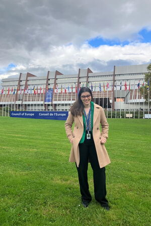 Autorin steht auf der Wiese vor dem Gebäude des Europarats. Im Hintergrund sieht man ein großes Schild wo „Council of Europe“ und „Conseil de l‘Europe“ steht. Das Gebäude des Europarats ist sehr breit und etwa 20 Meter hoch. Vor dem Gebäude und hinter der