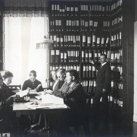 Schwarzweißfotografie: In einem Raum steht ein Mann an sehr hohen Bücherregalen, links davon sitzen 7 Männer und Frauen in historischer Kleidung und scheinen Punktschrift zu schreiben und zu lesen.