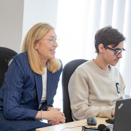 Am Schreibtisch sitzen zwei Personen. Die eine wird von der anderen am PC beraten.