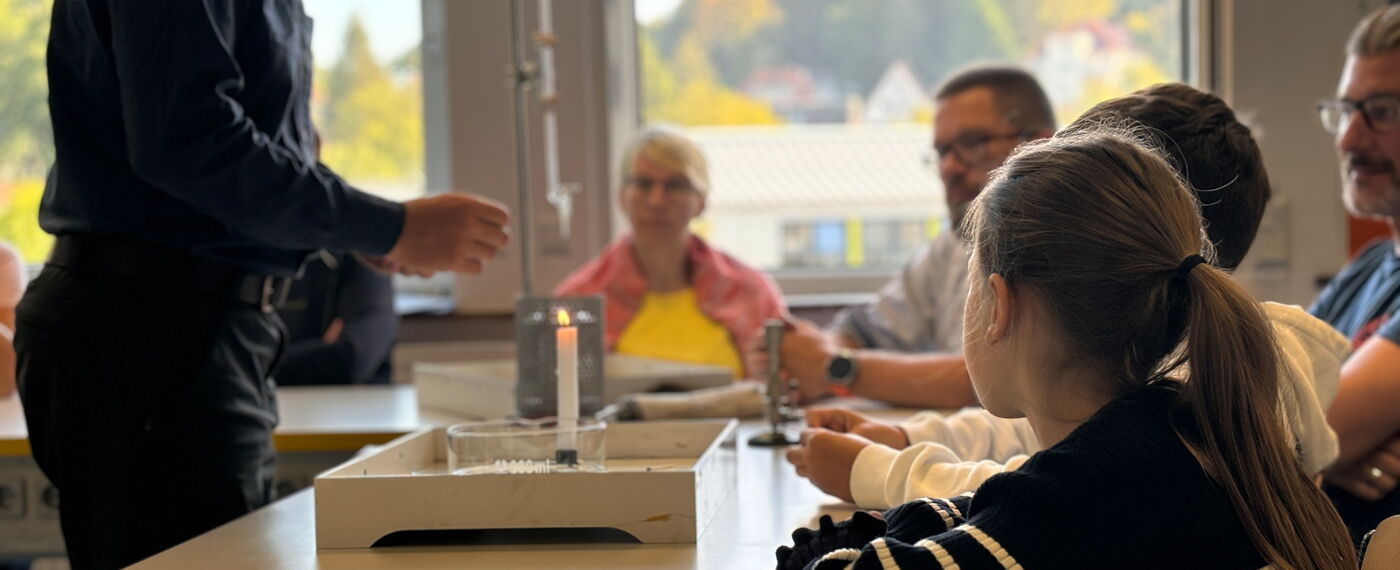Ein Lehrer demonstriert ein Experiment mit einer Kerze vor Schüler*innen und Lehrer*innen.