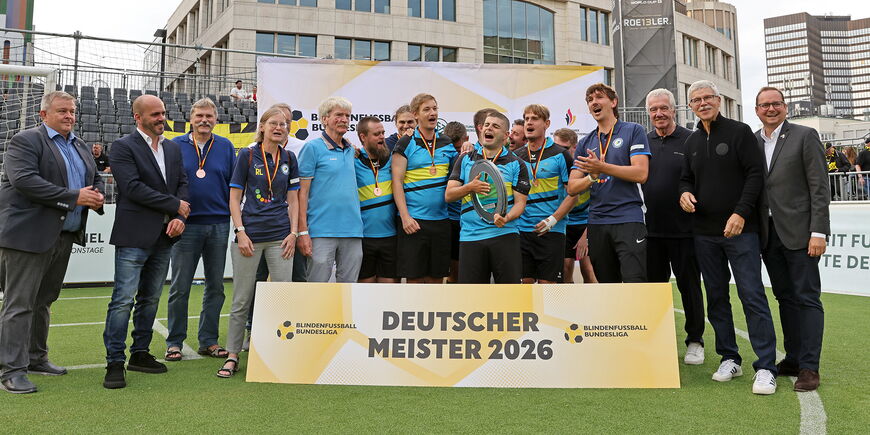 Das Team der SF BG blista steht jubelnd gemeinsam mit Menschen vom DFB auf dem Platz. vor ihnen ein großes Schild "Deutscher Meister 2026".