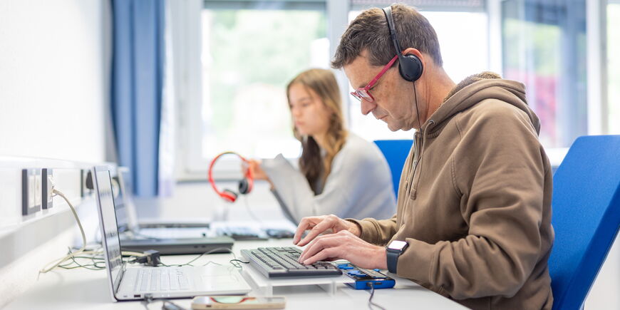 Zwei Teilnehmer sitzen in einem Unterrichtsraum an PCs und tragen Kopfhörer.