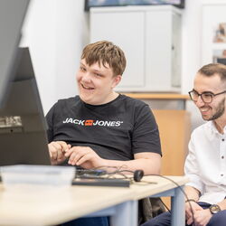 Zwei junge Männer sitzen gemeinsam an einem Pc und lächeln.