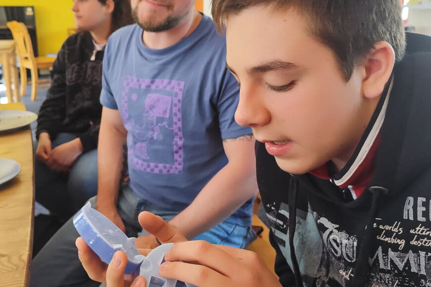 Ein blinder Schüler hat eine Gussform für einen Würfel in der Hand, ein Lehrer im Hintergrund lächelt in die Kamera.