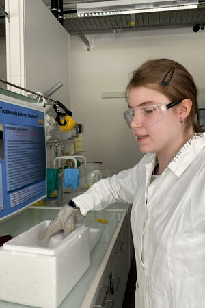 Lia hantiert im Chemielabor mit Trockeneis – sie trägt einen Laborhandschuh, einen weißen Laborkittel und eine Schutzbrille.