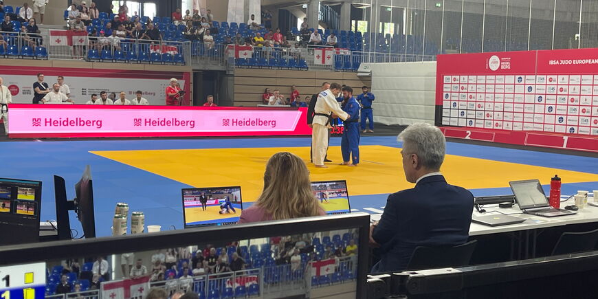 Blick über die Schulter der Juror*innen auf Bildschirme bei der Para-Judo-Europameisterschaft.  Zwei Judoka kämpfen.