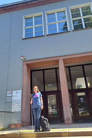 Lia auf der Treppe vor dem Fachbereich Chemie der Uni Marburg – sie hält ihren Langstock in der Hand und hat einen Rucksack neben sich.