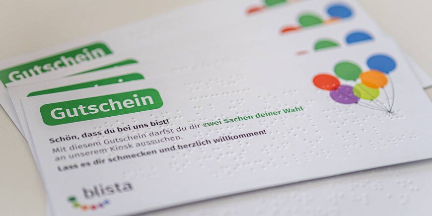 Nahaufnahme eines Gutscheins für neue Schüler*innen, darauf sind Punktschrift, bunte Ballons und das blista-Logo zu erkennen.