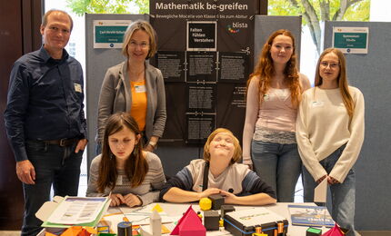 Tobias Mahnke und Maike Castorph stehen mit 4 Schüler*innen an einem Messestand, hinter ihnen ein schwarzes Plakat mit blista-Logo, vor ihnen verschiedene bunte Tastmodelle.