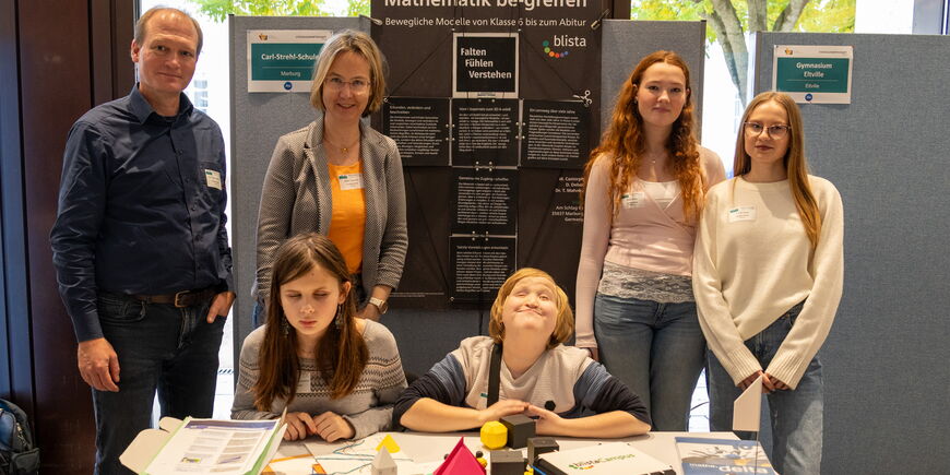 Tobias Mahnke und Maike Castorph stehen mit 4 Schüler*innen an einem Messestand, hinter ihnen ein schwarzes Plakat mit blista-Logo, vor ihnen verschiedene bunte Tastmodelle.