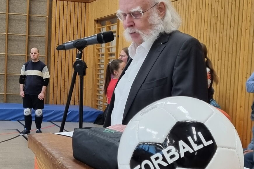 Jürgen Hertlein steht in der blista-Sporthalle vor einem Mikrofon, vor ihm liegt ein Torball.