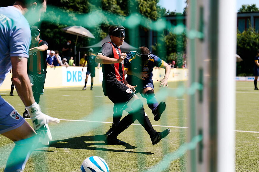 Blick durch das Tornetz beim Blindenfußball, zwei Spieler haben gerade einen Ball geschossen, den der Torwart versucht zu fangen.