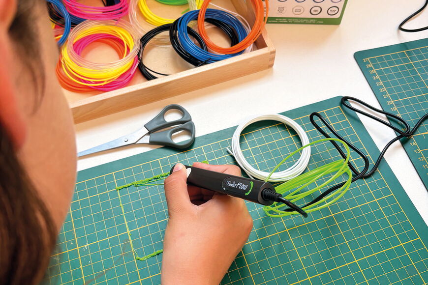 Blick über die Schulter einer Schülerin: sie hat einen 3-D-Druck-Stifit in der Hand und erzeugt damit auf einer grünen Bastelunterlage ein quadratisches Gebilde. Daneben liegt buntes Filament.