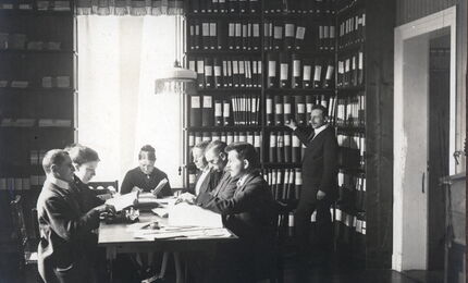 Schwarzweißfotografie: In einem Raum steht ein Mann an sehr hohen Bücherregalen, links davon sitzen 7 Männer und Frauen in historischer Kleidung und scheinen Punktschrift zu schreiben und zu lesen.