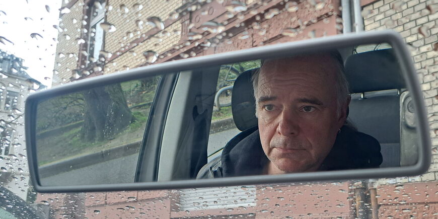 Herr W ist im Rückspiegel eines Autos zu sehen, auf dessen Windschutzscheibe Regentropfen die Sicht auf ein Haus verdecken.