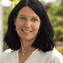 Portraitfoto Susanne Becker - sie hat dunkles schulterlanges Haar, trägt eine rahmenlose Brille und eine weiße Bluse.