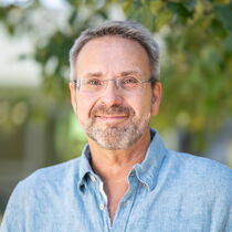 Portraitfoto Carsten Erbes - Er trägt kurzes graues Haar, einen Bart und eine Brille und hat ein hellblaues Hemd an.