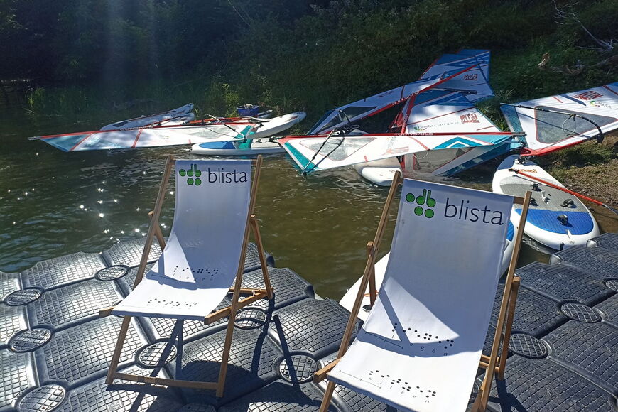 Auf einem Ponton auf einem Gewässer stehen blista-Liegestühle, dahinter liegen Windsurfbretter im Wasser.