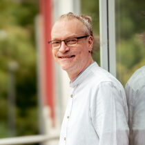 Portraitfoto Frank Stollenwerk - er trägt ein weißes Hemd, eine Brille und hat das lange Haar zu einem Dutt gebunden.