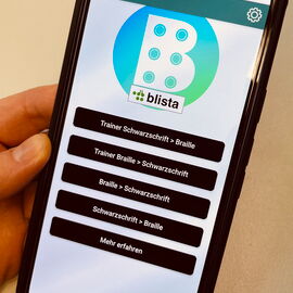 Eine Hand hält ein Smartphone, auf dem die Braille-Trainer App zu sehen ist.