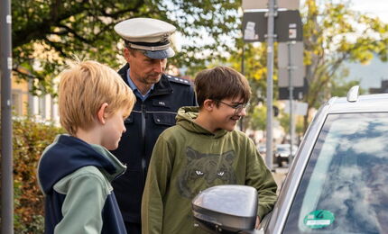 Zwei Schüler und ein Polizist stehen neben einem Auto am Straßenrand und reden mit einer Person im Auto.