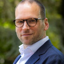 Portraitfoto Mirko Melz - er trägt Brille, Dreitagebart, weißes Hemd und blaues Sakko.
