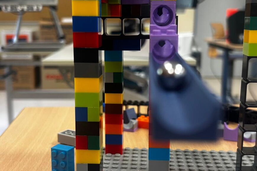 In einem Klassenraum ist eine Kugelbahn aus Lego Duplo Steinen mit hohen Türmen aufgebaut. Eine Metallkugel rollt auf die Kamera zu.