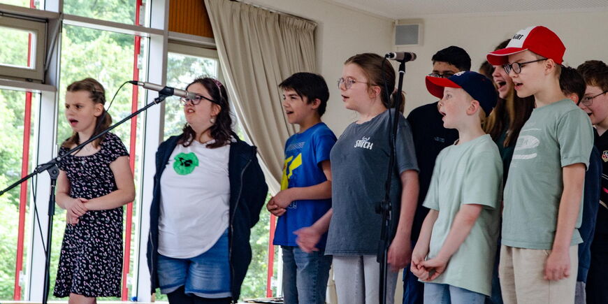 Eine Gruppe Kinder in sommerlicher Kleidung steht auf einer Bühne und sing die Standmikrofone.