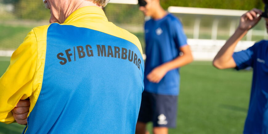 Während des Fußballtrainings sieht man den Rücken des Trainers mit Trainingsjacke im Vordergrund, während die Spieler im Hintergrund trainieren.