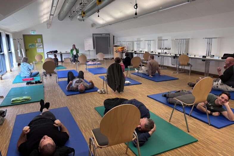 Eine bunte Gruppe aus Mitarbeitenden liegt oder sitzt auf Matten und führt Entspannungsübungen aus.