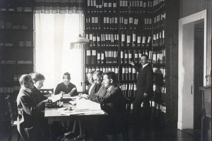 Schwarzweißfotografie: In einem Raum steht ein Mann an sehr hohen Bücherregalen, links davon sitzen 7 Männer und Frauen in historischer Kleidung und scheinen Punktschrift zu schreiben und zu lesen.