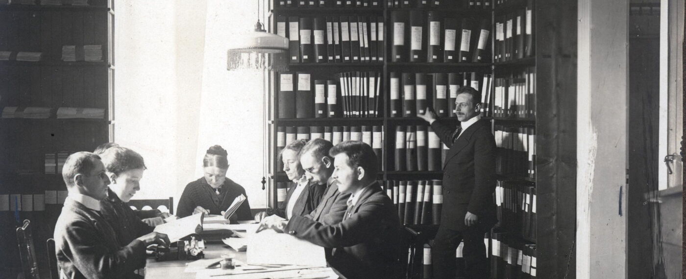 Schwarzweißfotografie: In einem Raum steht ein Mann an sehr hohen Bücherregalen, links davon sitzen 7 Männer und Frauen in historischer Kleidung und scheinen Punktschrift zu schreiben und zu lesen.