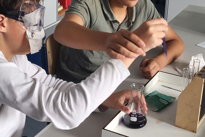 Zwei Schüler sitzen vor einem Tisch, vor ihnen steht ein Experimentiertablett. Auf dem Tablett steht ein Reagenzglasständer mit Reagenzgläsern, ein Erlenmeyerkolben mit einer lila Flüssigkeit sowie eine Tüte Kaisernatron. Ein Schüler hat eine Augenbinde a