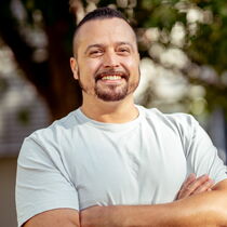 Portraitfoto Janosch Briel - er trägt kurzes dunkles Haar, einen Bart und ein weißes T-Shirt.