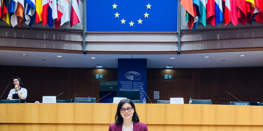 Autorin im EU Parlaments Sitzungssaal. Über dem Kopf der Autorin sieht man ein Schild mit einem Kreis aus Sternen, das Symbol der EU. Links und rechts davon hängen die Flaggen aller 27 EU-Mitgliedsländer. Unter dem Schild steht der Begriff „Europäisches P