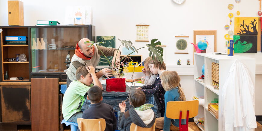 Ein Lernbegleiter demonstriert einer Gruppe Schüler*innen etwas an einer Pflanze