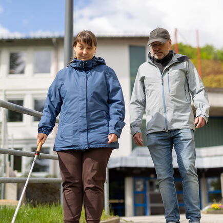 Eine Frau mit einem Langstock steht an einem Treppengeländer, ein Lehrer für O&M steht neben ihr.