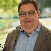 Thorsten Büchner trägt ein hellblaues Hemd, ein braunes Sakko und eine Brille. Er hat braunes Haar und einen Langstock in der Hand.