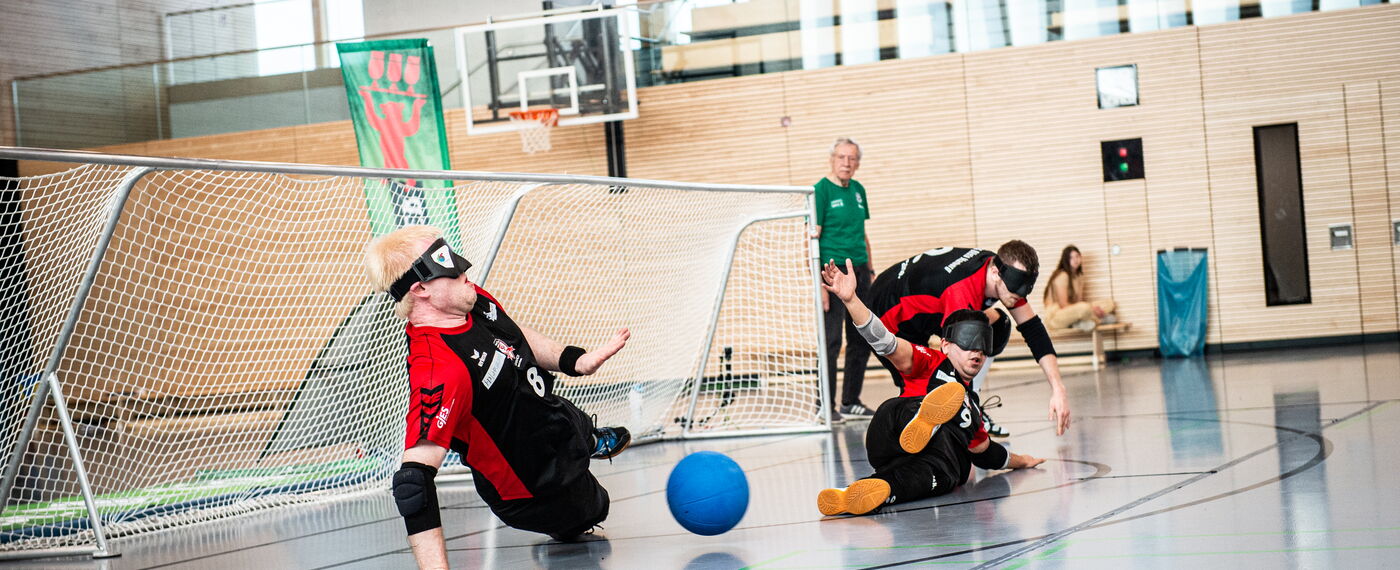 In einer Sporthalle werfen sich drei Goalballspieler vor ein Tor, um einen Ball abzuwehren.