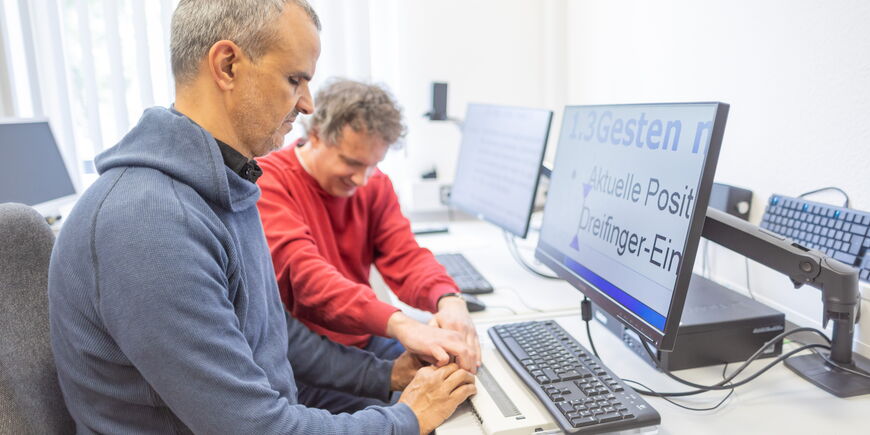 Zwei Männer sitzen an einem PC und haben die Hände gemeinsam an einer Braillezeile.