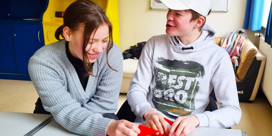 Eine Schülerin und ein Schüler sitzen in einem Klassenraum und puzzeln mit dreieckigen Teilen eines MuLi-Lernpaketes.