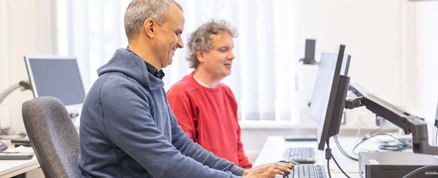 Zwei Männer sitzen in einem hellen Raum an einem Computer.