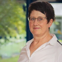 Portraitfoto Irene Noll - sie hat kurze braune Haare, eine Brille und trägt eine weiße Bluse.