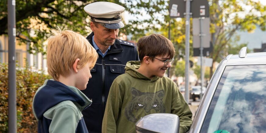 Zwei Schüler und ein Polizist stehen neben einem Auto am Straßenrand und reden mit einer Person im Auto.